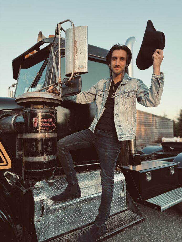 Male performer from The Wild Ones Country Band posing beside a classic black truck with a cowboy hat